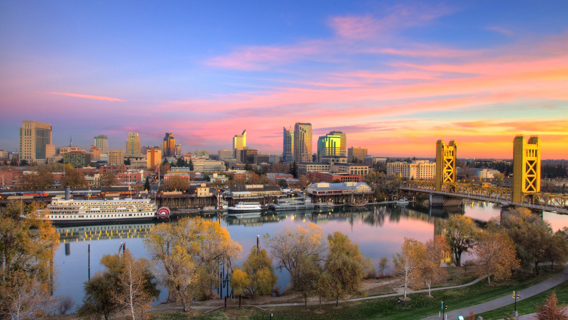 Panoramic view of downtown Sacramento at sunset, showing the city skyline, the Tower Bridge with its golden towers, and the Sacramento River with boats docked along the waterfront