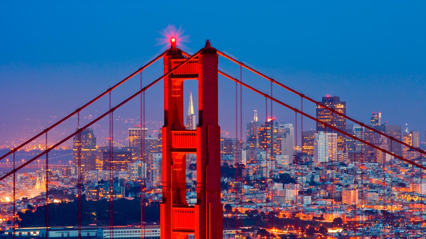 Golden Gate Bridge at dusk with its red towers illuminated, overlooking the San Francisco skyline with city lights glowing against a deep blue sky
